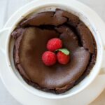 Tall espresso-infused chocolate soufflé in ramekin dusted with powdered sugar, topped with fresh raspberries, mint, and whipped cream-dramatic airy dessert