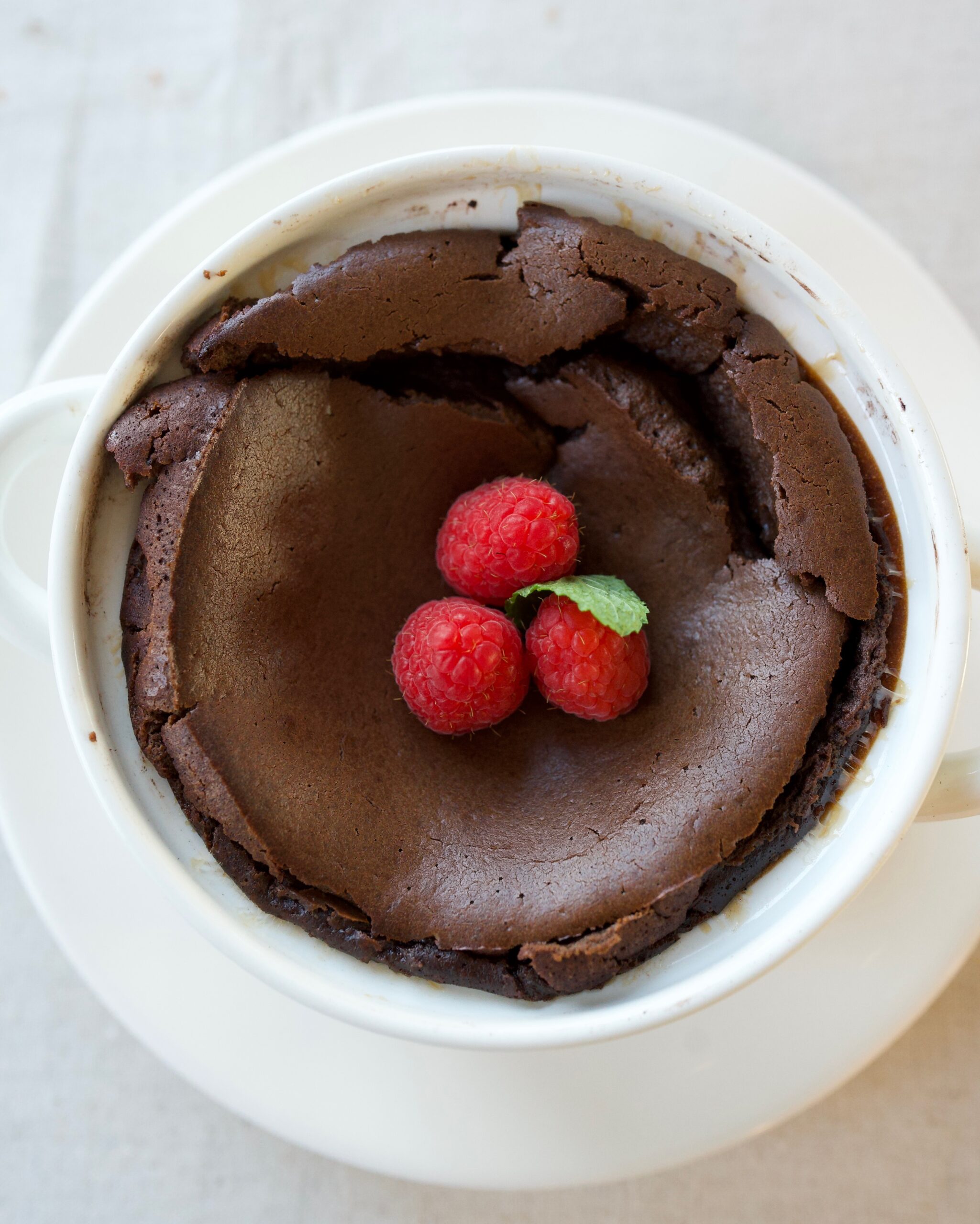 Tall espresso-infused chocolate soufflé in ramekin dusted with powdered sugar, topped with fresh raspberries, mint, and whipped cream-dramatic airy dessert